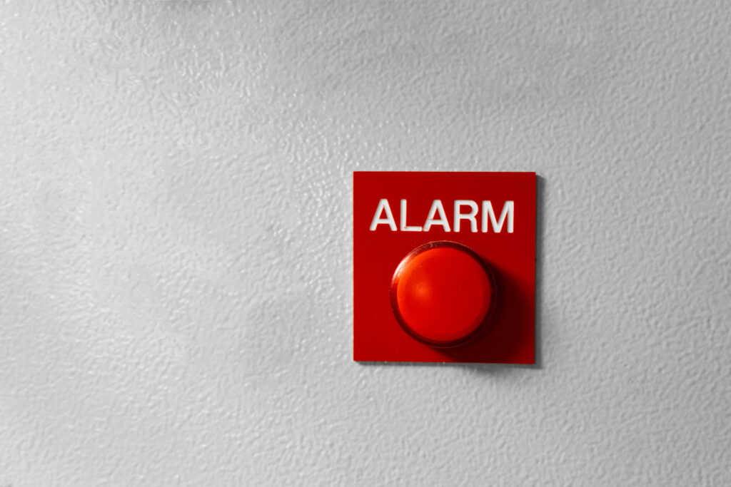 A hardwired panic alarm mounted on a grey wall for easy activation by anyone who spots a threatening situation.